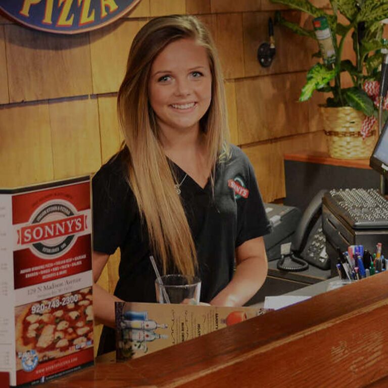 Smiling woman stands at Sonny’s Italian Kitchen & Pizzeria counter with menu and cash register.